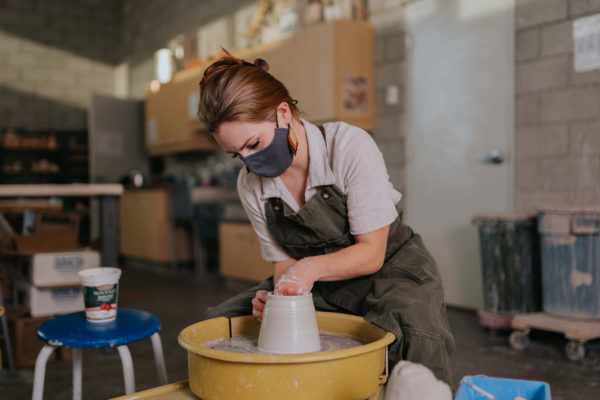 Red haired woman wearing a face mask throws a clay vessel on the pottery wheel