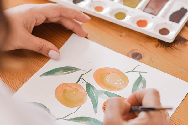 Woman paints a watercolour of oranges on paper with a palette in the background