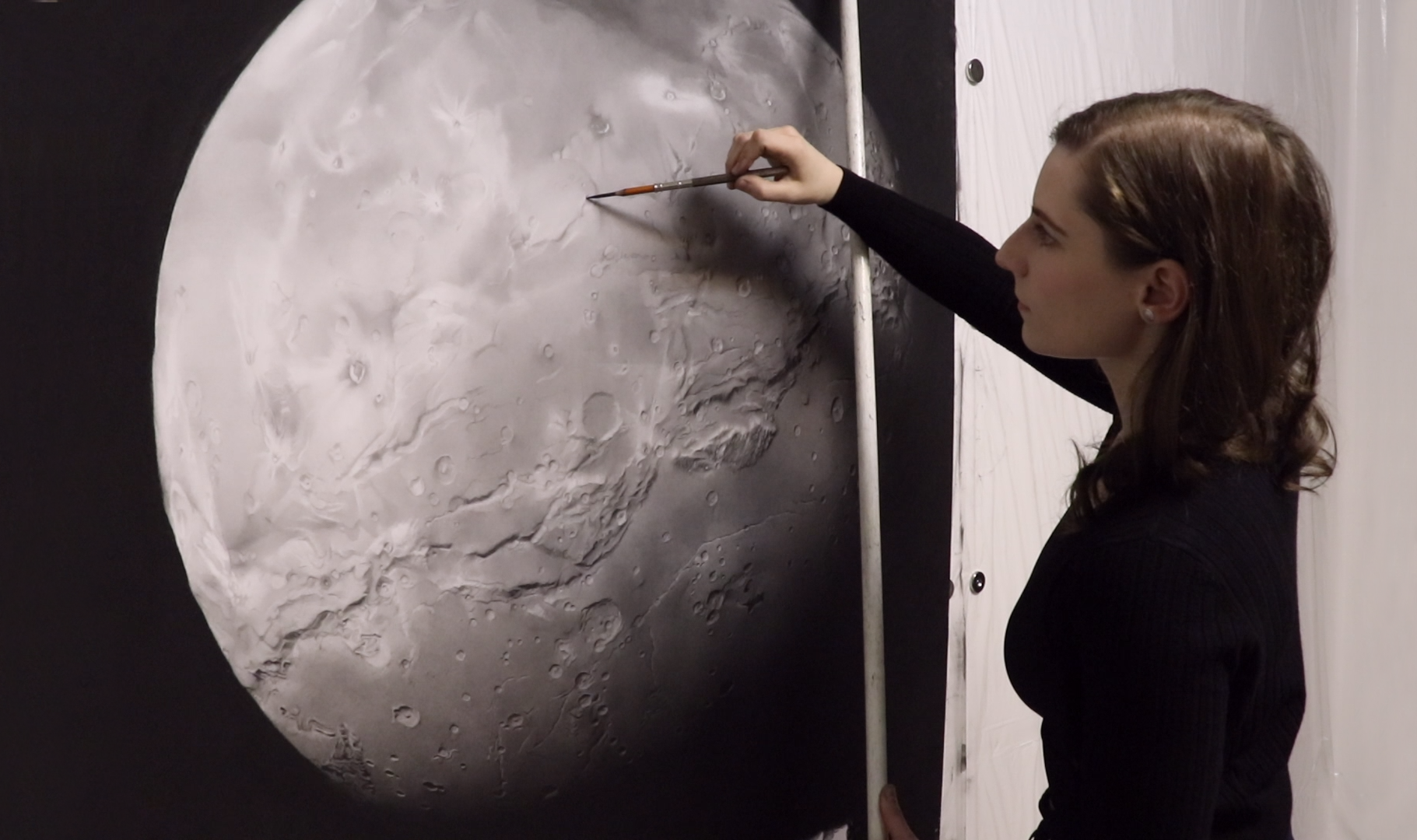 MArina Fridman drawing with a pencil in front of a moon drawing on the wall.