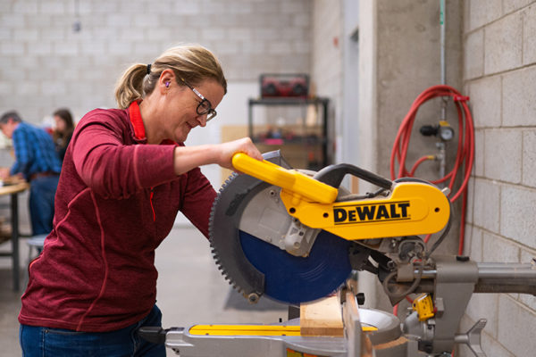 A woman uses a mitre saw in a wood shop to cut down a piece of 2 by 4 wood.