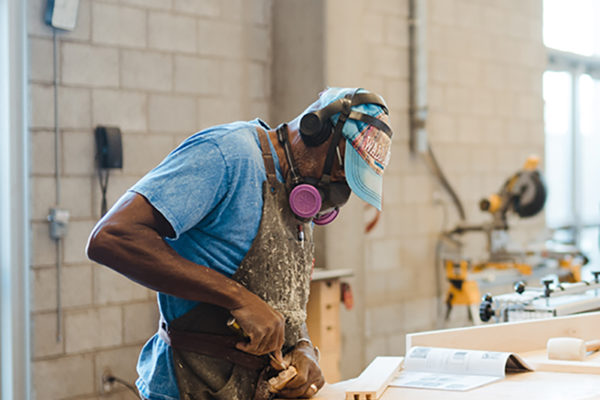 A person wearing a blue hat, safety googles, a respirator and a grey apron is working on joinery in the Wood Studio.