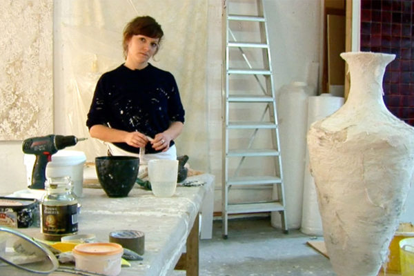 A woman stares at the camera while working in her studio. A large ceramic vessel sits in the foreground.