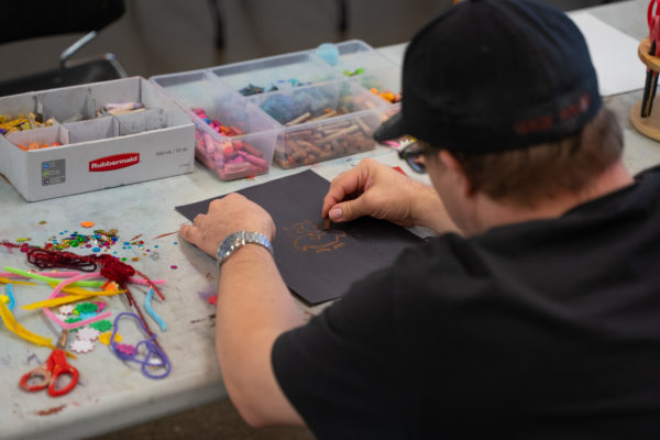 and over the shoulder view of a man in a black tshirt and black baseball cap drawing an owl onto a black piece of paper. an assortment of art supplies are next to him on the table