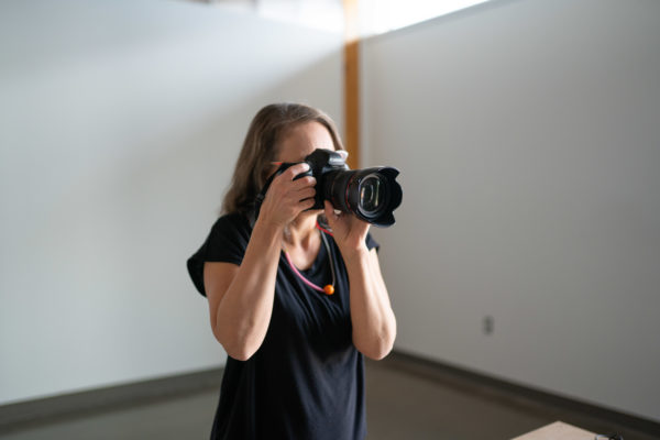 Woman wearing black clothing taking a picture with a digital camera in a white classroom