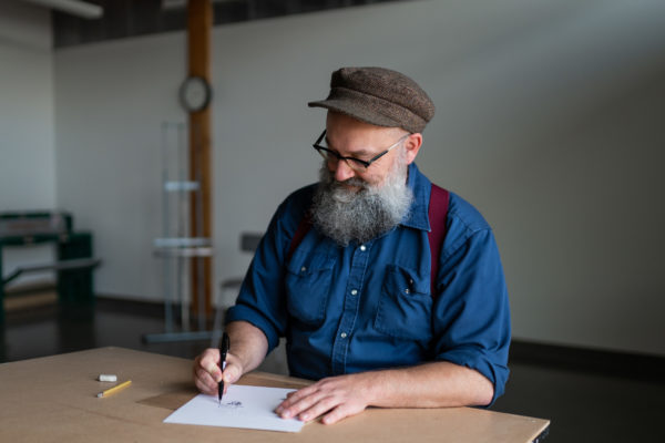A man wearing a hat, glasses and a blue long sleeve button up shirt with red suspenders is drawing a comic. He has a long gray beard.