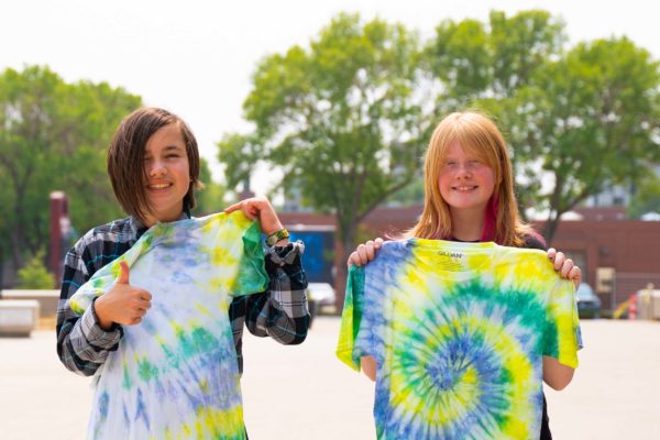 two youth stand outsdie holding tie-dye shirts, one of them is motioning a thumbs up sign