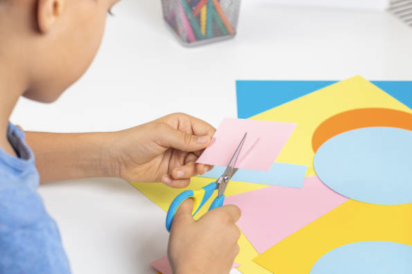 a close up view of a child cutting a pink piece of paper, there are yellow and blue pieces of cut up paper on the table